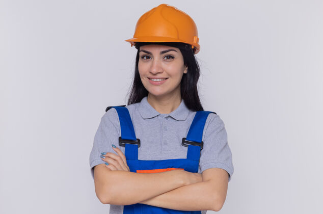 女人身穿施工制服 头戴安全帽的年轻建筑工人站在白色的墙上 面带自信的微笑 双臂交叉建筑手臂制服