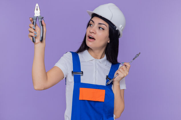 尝试身穿施工制服 头戴安全帽 手持扳手和钳子的年轻建筑工人站在紫色的墙上 困惑地试图做出选择建筑商困惑头盔