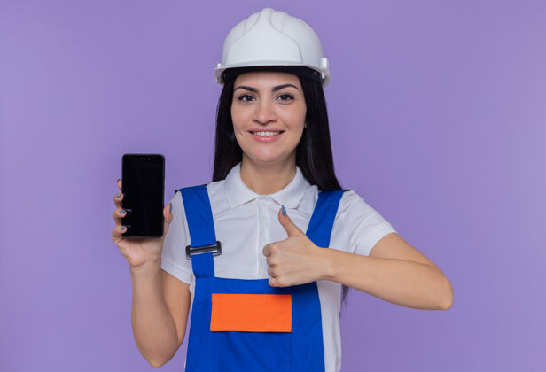 年轻身穿建筑制服 头戴安全帽的年轻建筑工人展示着智能手机 面带微笑 自信地竖起大拇指站在紫色的墙上展示女人站立
