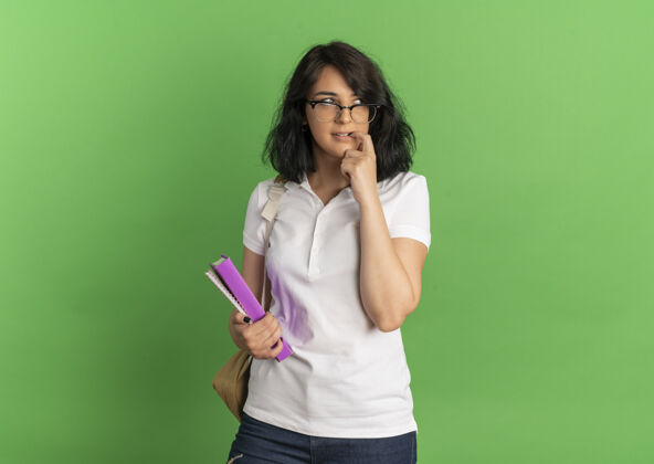 穿年轻焦急漂亮的高加索女学生戴着眼镜背着书包咬着手指拿着书放在绿色的复印空间里抱复制焦虑