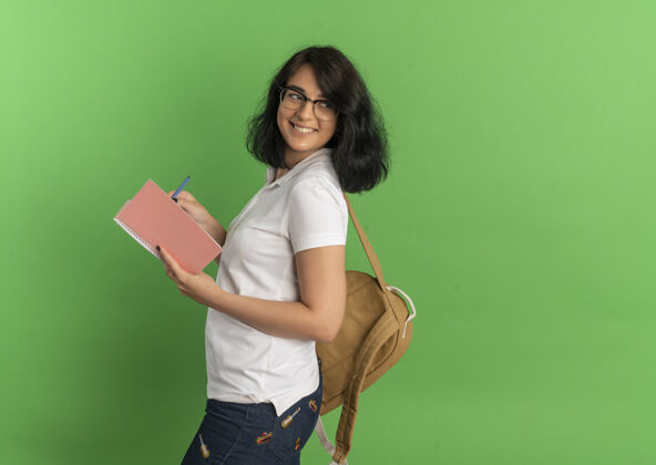 抱年轻的高加索女孩 面带微笑 戴着眼镜 背着书包 站在一旁 手里拿着笔和笔记本 放在绿色的空白处背空间眼镜
