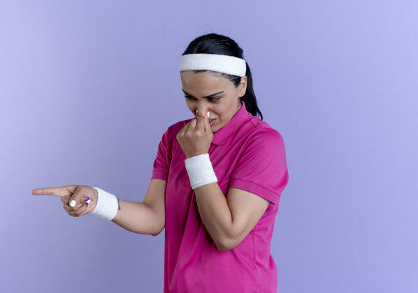 鼻子年轻恼怒的白人运动女性戴着头带和腕带闭着鼻子指着紫色的侧面和复制空间关闭运动腕带