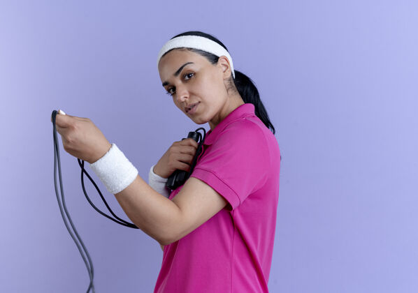 空间年轻自信的白人运动女性戴着头带和腕带站在一边拿着跳绳看着相机紫色的复制空间立场头带绳子