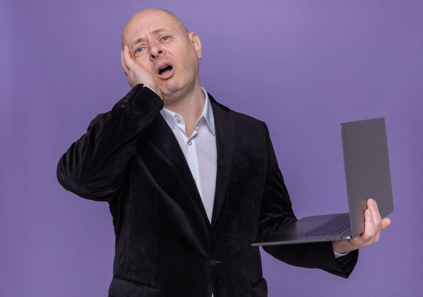 困惑中年光头西装革履 手持笔记本电脑 看在前面 手放在头上 误站在紫色的墙上错了西装拿着