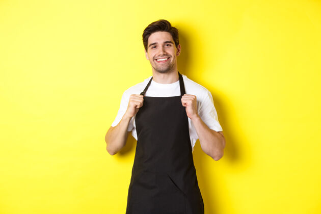 黄色自信的咖啡师穿着黑色围裙站在黄色背景下服务员微笑着 看起来很高兴自信厨师烹饪