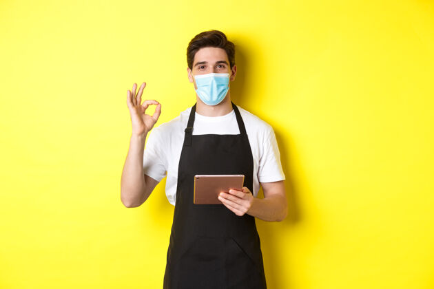 面膜covid-19的概念 小型企业和流行病戴着医用面罩和黑色围裙的销售员显示ok标志 用黄色背景的数字平板电脑接受订单男人黄色时尚