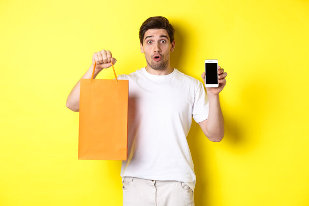 站着惊讶的男人展示手机屏幕和购物袋 站在黄色背景下复制空间工作室男人表情