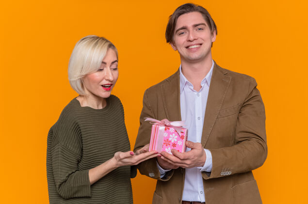 在一起年轻漂亮的情侣快乐的男人站在橘色的墙上给他可爱的微笑的女友一份礼物一起欢庆国际妇女节爱女人礼物