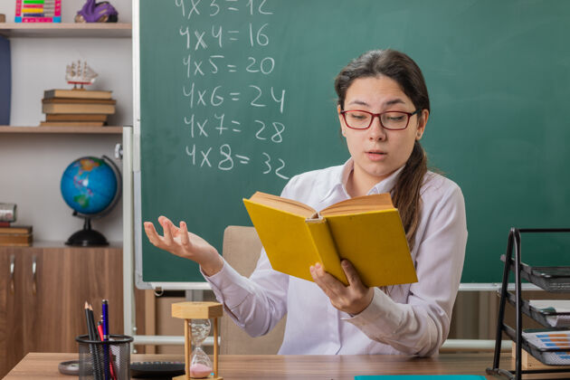 课戴眼镜的年轻女老师拿着备课用书坐在教室黑板前的课桌旁教室书桌穿