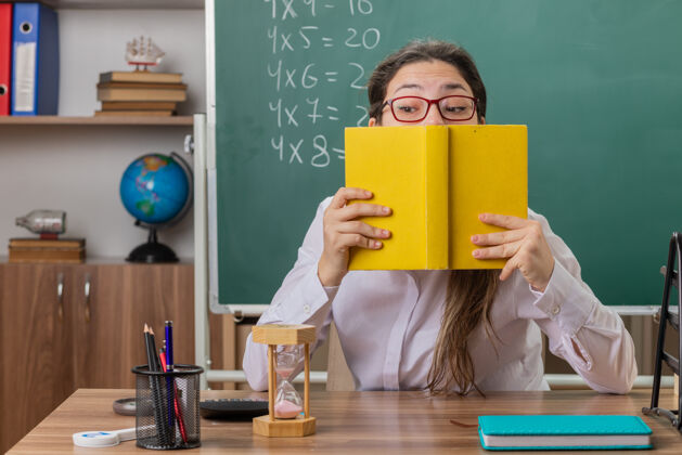 老师戴眼镜拿着备课书的年轻女教师坐在教室黑板前的课桌旁 困惑而焦虑前面教室年轻