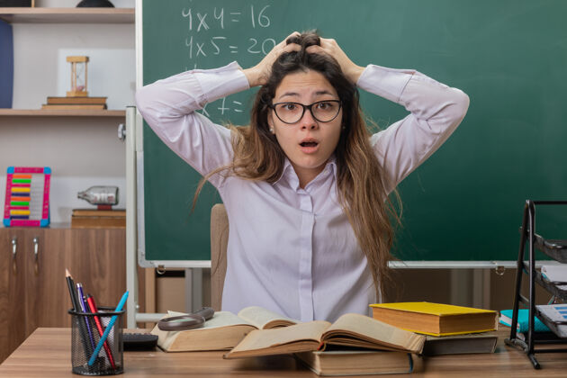 担心年轻的女老师戴着眼镜 拿着书 手放在头上 坐在教室黑板前的课桌旁 神情迷茫 忧心忡忡眼镜困惑教室