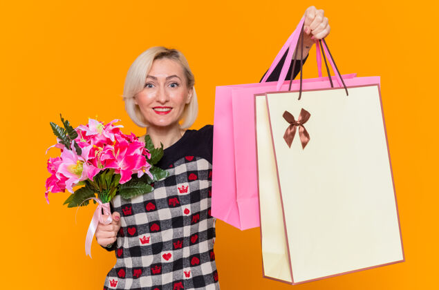 女人身着漂亮衣服的快乐年轻女子手持鲜花和礼品纸袋 站在橙色的墙上 微笑着欢庆国际妇女节快乐微笑衣服
