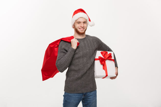 胡须年轻的快乐胡子男人拿着圣诞老人包和白色礼物大礼品盒圣诞节