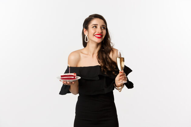 黑发庆祝和派对概念时尚的女人拿着生日蛋糕和蜡烛 喝着香槟 微笑着看着一边 站在白色的背景上年轻女人生日