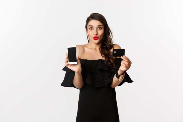 时尚时尚和网上购物概念快乐的年轻女子穿着黑色连衣裙 展示信用卡和手机屏幕 站在白色背景上网络营销年轻美丽