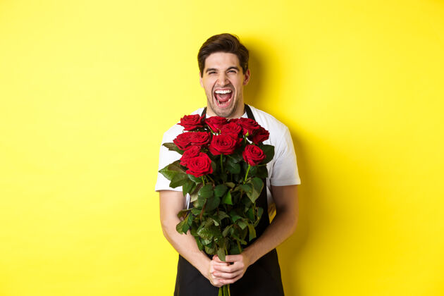 花店穿着黑色围裙 手持玫瑰花束的有趣的年轻推销员 站在黄色背景下的花店老板哈哈大笑工作室帅哥男人