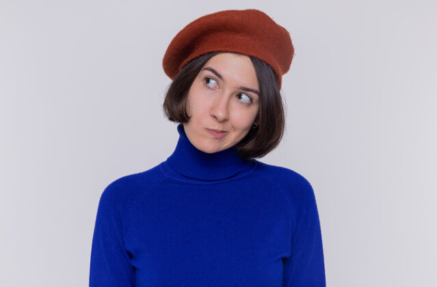 年轻身穿蓝色高领毛衣 头戴贝雷帽 留着短发的年轻女子站在白色的墙上 带着怀疑的表情向一旁望去站着贝雷帽头发