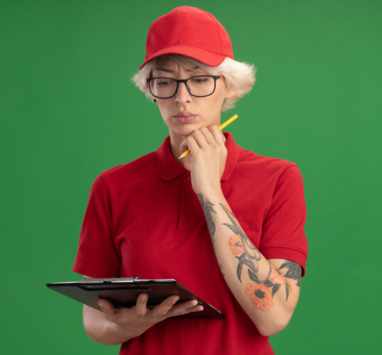 穿身穿红色制服 戴着帽子的年轻送货员戴着一副眼镜 上面有空白页和铅笔的剪贴板 站在绿色的墙壁上 面带严肃的思考站眼镜交货