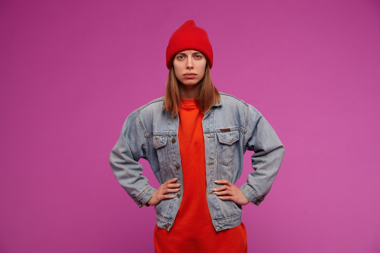 欧洲人年轻女子的肖像 深色长发 穿着牛仔裤夹克 红色毛衣和帽子 手放在腰上 皱着眉头隔着紫色的墙青少年皮肤漂亮