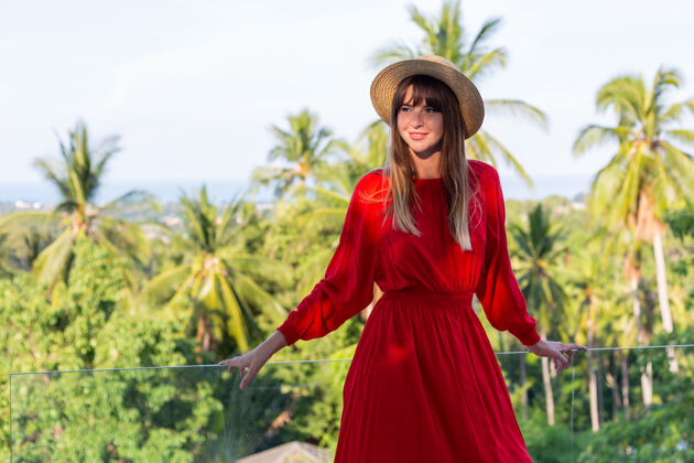 沙滩穿着红色夏装 戴着草帽 在阳台上享受热带海景和棕榈树的快乐女人放松女士年轻
