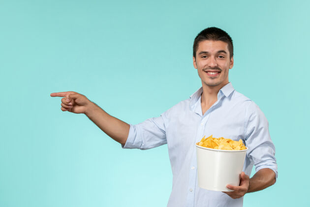 孤独正面图：年轻的男性拿着篮子和薯片 在蓝色的表面上微笑着看电影实验服远程看
