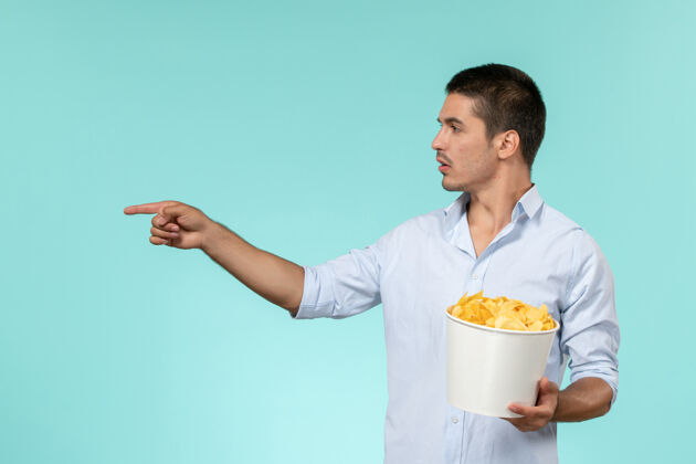 服务员正面图：年轻的男性拿着篮子和薯片 在浅蓝色的表面上看电影远程年轻的男篮子
