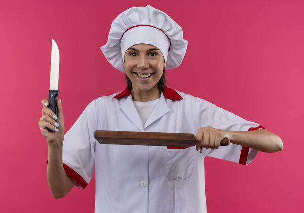 年轻年轻快乐的白人厨师女孩穿着厨师制服 拿着刀和砧板隔离在粉色空间和复印空间制服举行粉红