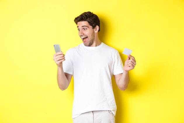 工作室惊讶的家伙拿着智能手机和信用卡 在黑色星期五网上购物 站在黄色背景下时尚成年人表情
