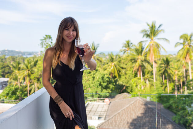 放松在热带阳台上 一个穿着黑色晚礼服 端着一杯葡萄酒的女人树平静黑色