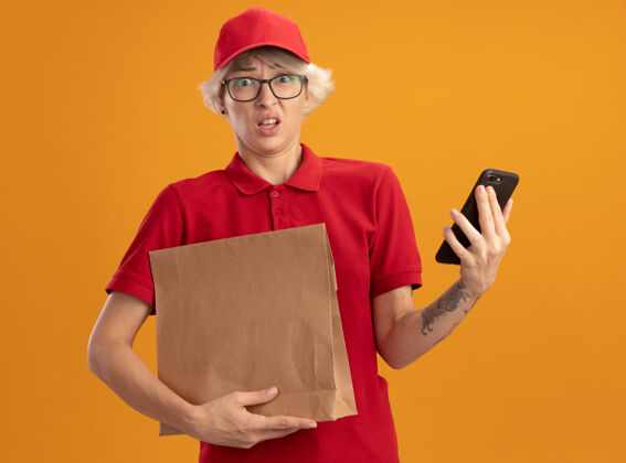 送货身穿红色制服 戴着帽子 拿着纸包和智能手机的年轻女送货员站在橙色的墙上 感到困惑和焦虑焦虑帽子移动