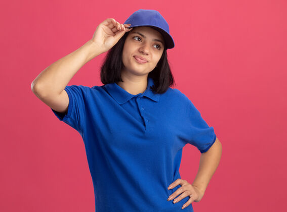 帽子不满意的年轻送货女孩 穿着蓝色制服 戴着帽子 站在粉红色的墙上困惑地看着一边靠边站制服拼图