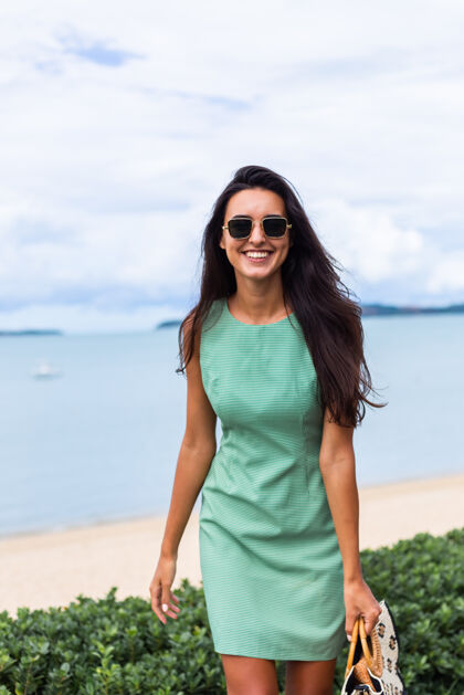 时尚漂亮时尚的快乐女人 穿着绿色夏装 带着包 度假时戴着太阳镜 背景是蓝色的大海海滩热带度假