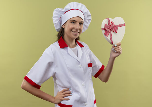 女孩身着厨师制服 面带微笑的白人年轻厨师女孩手持心形盒子 将手放在腰上 隔离在绿色空间和复制空间放着手形状