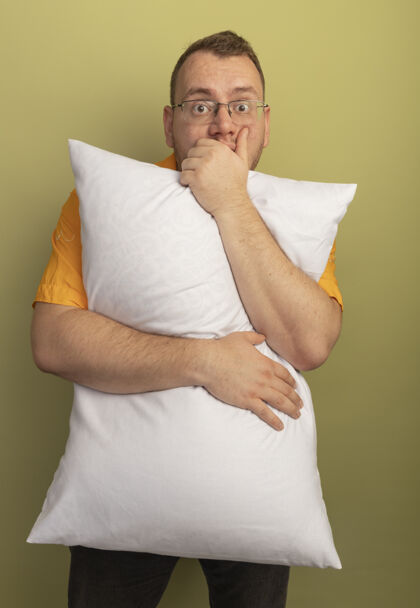枕头戴着眼镜的男人穿着橘色衬衫抱着枕头惊讶地捂着嘴用手站在轻墙上手站着穿上