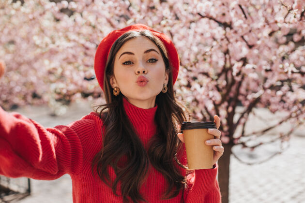 花快乐的女人手里拿着一杯茶吹着吻自拍穿着红毛衣的女人拿着咖啡杯对着盛开的樱花的画像季节肖像无忧无虑