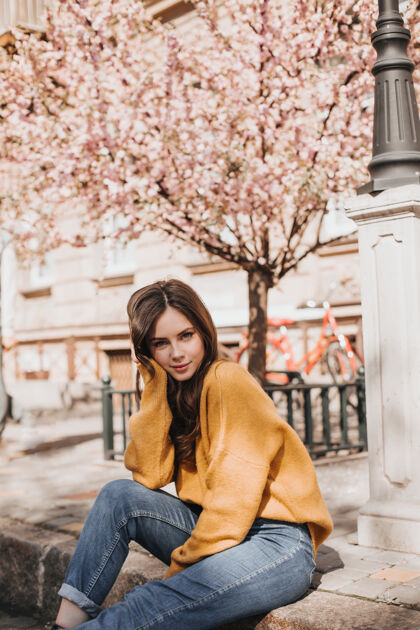 自由穿着橘色毛衣的女人坐在马路上穿着牛仔裤的迷人女孩在樱花附近摆姿势女人看着镜头太阳镜花衣服