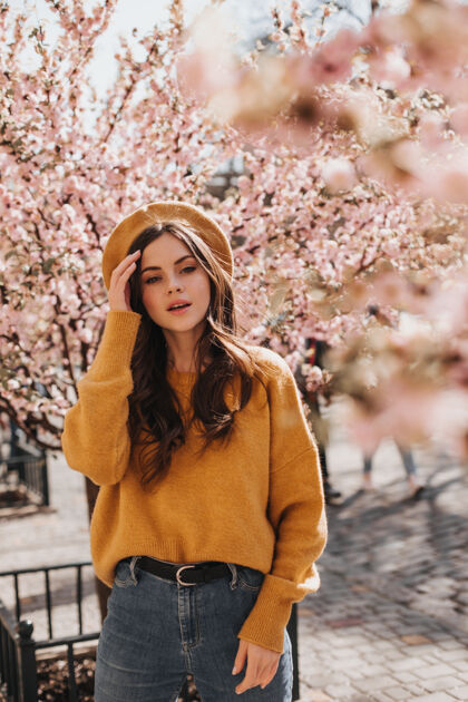 健康身着时髦服装 头戴帽子的黑发女孩在樱花附近摆姿势身着橙色毛衣 牛仔裤和贝雷帽的女人在盛开的花园里散步的肖像温柔太阳镜自然
