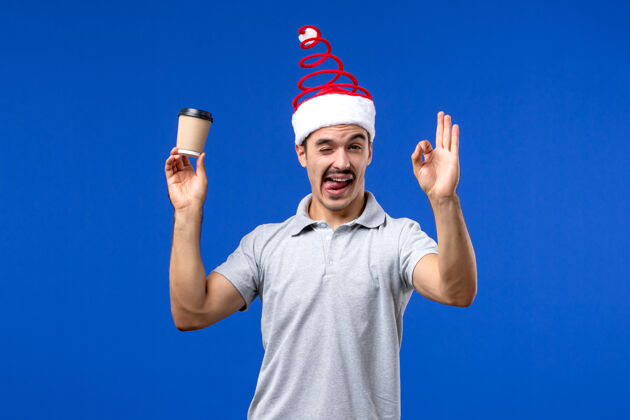 人正面图：年轻男性拿着塑料咖啡杯在蓝色办公桌上新年男性假期桌子塑料帽子
