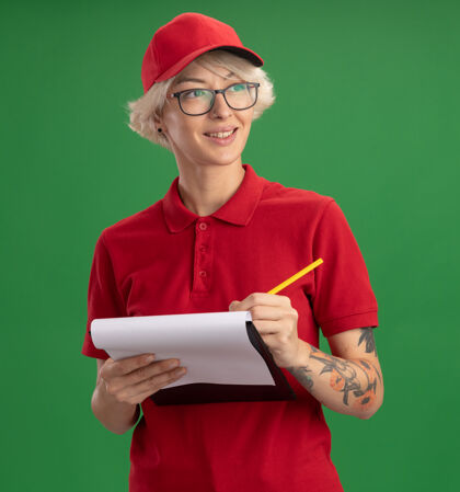 眼镜年轻的送货员身穿红色制服 戴着一顶帽子 戴着一副眼镜 上面有空白页和铅笔 脸上带着微笑 站在绿色的墙上写着什么剪贴板帽子铅笔