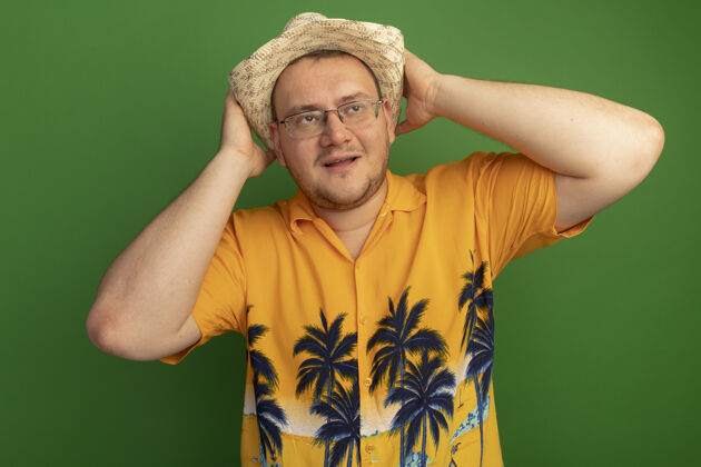 帽子一个戴着眼镜的男人 穿着橘色衬衫 戴着夏天的帽子 脸上挂着幸福的表情 头后站在绿色的墙上男人脸站