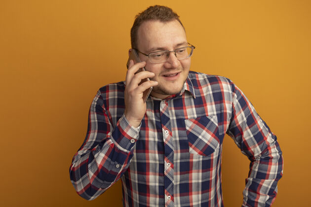 男人一个戴着眼镜和格子衬衫的男人站在橘色的墙上一边用手机说话一边微笑着微笑站立同时