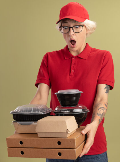 站着年轻的送货员身穿红色制服 戴着眼镜 手里拿着比萨饼盒和食品包 站在绿色的墙上 惊讶地看着它们食物帽子惊喜