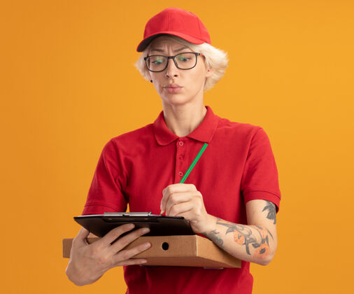 制服身穿红色制服 戴着帽子 戴着眼镜 拿着披萨盒和铅笔的剪贴板 站在橙色的墙上 严肃地写着什么剪贴板拿着脸