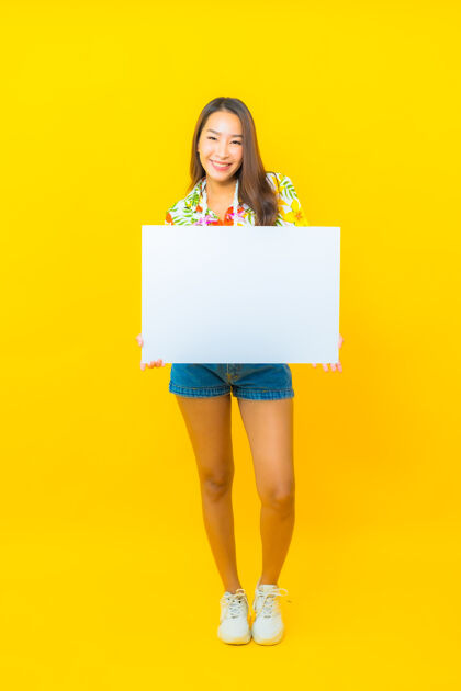 女孩美丽的亚洲年轻女子的画像 黄色墙上挂着白色的空广告牌微笑模型举行