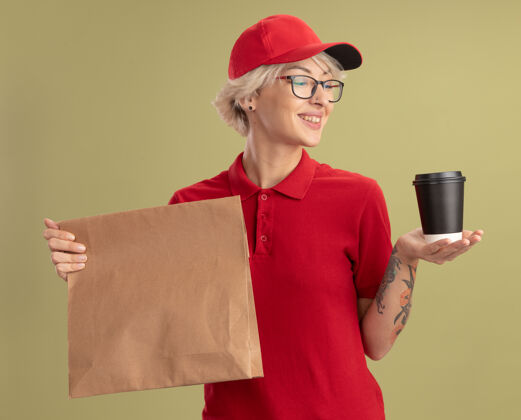 年轻人年轻的送货员身穿红色制服 戴着帽子 戴着眼镜 手里拿着纸包和纸杯 站在绿色的墙边自信地微笑着自信抱着站着