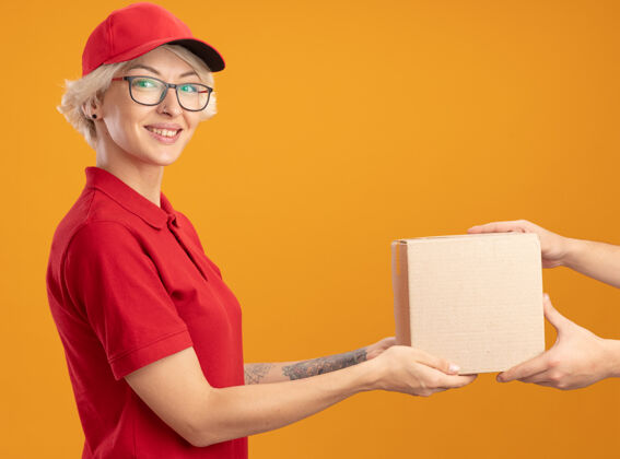 送货身着红色制服 戴着眼镜的年轻送货员站在橙色的墙上 友好地微笑着给你的顾客送纸盒穿朋友眼镜