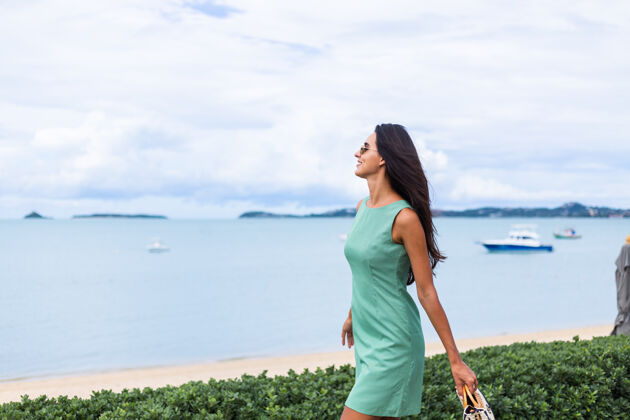 海滩漂亮时尚的快乐女人 穿着绿色夏装 带着包 度假时戴着太阳镜 背景是蓝色的大海休闲年轻时尚