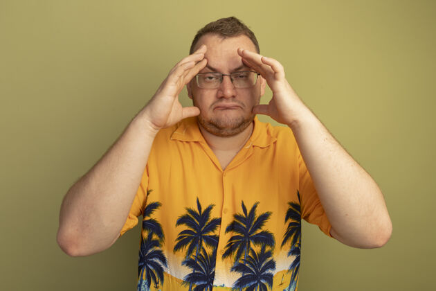 不舒服一个戴着眼镜 穿着橘色衬衫的男人站在绿色的墙上 看起来不舒服 摸着头 头痛得厉害站着摸着头