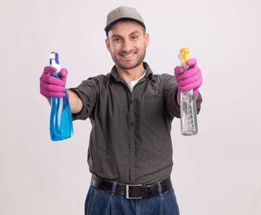 喷雾年轻的清洁工穿着休闲服 戴着橡胶手套 手里拿着喷雾瓶 站在白色的墙上 面带微笑帽子立场橡胶