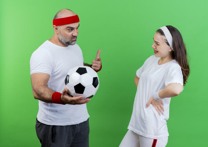 情侣一对戴着头带和腕带的运动型成年夫妇自信的男人拿着足球 指着足球 让女人印象深刻 手放在腰上 在绿色的墙上互相孤立地看着对方女人穿指向